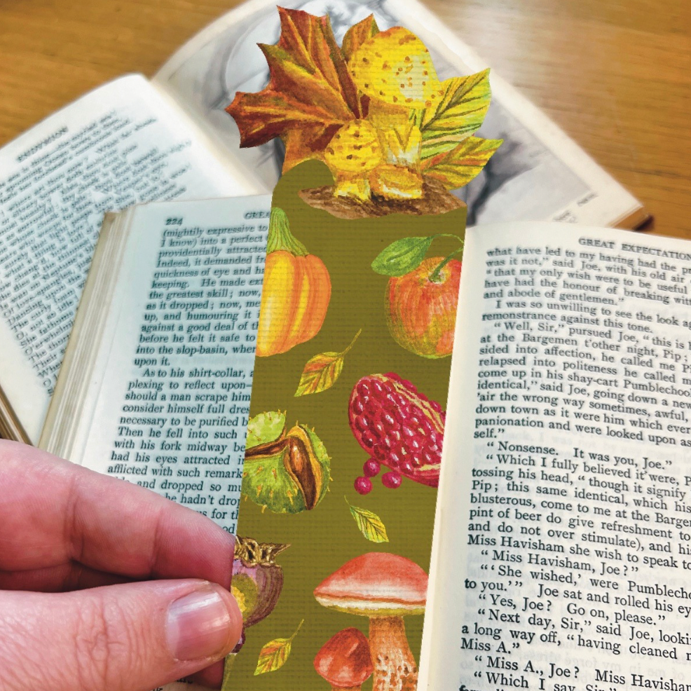 Bookmark in moss green illustrated with harvest fruits and mushrooms, die cut to the shape of maple leaf and mushrooms. Bookmark is held ion front of the open pages of a book.