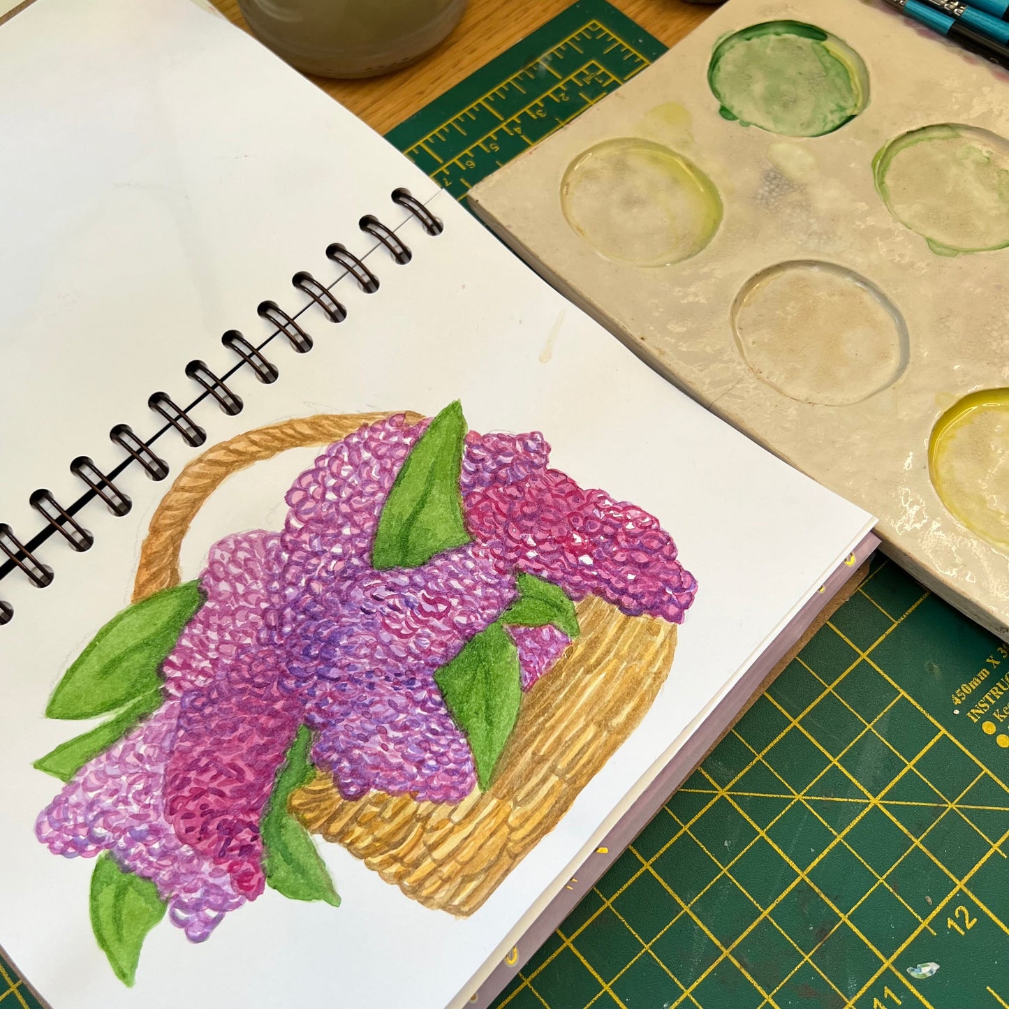 Watercolour illustration of a bracket of lilacs in a sketchbook page, on a wooden desk with a green craft board and paint palette