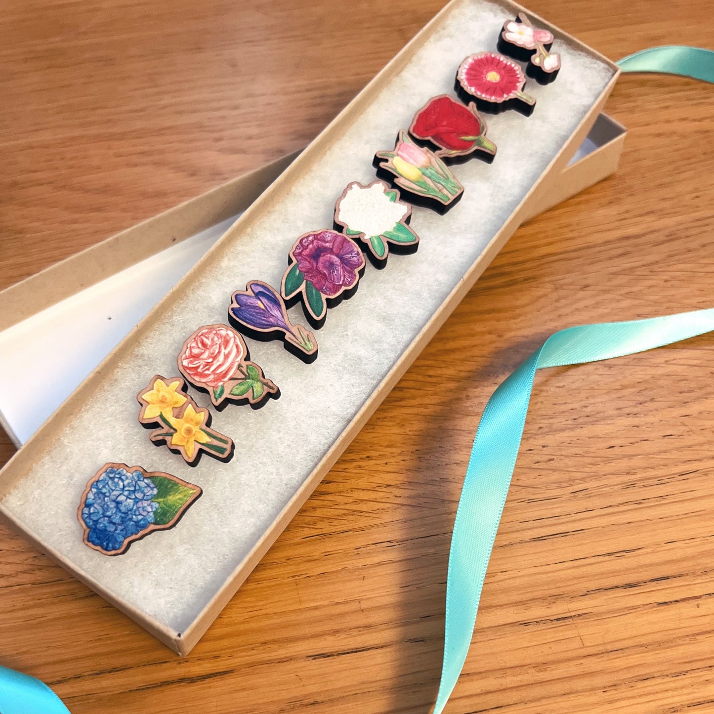 Gift set of 10 floral wooden pins presented in a watch jewellery gift box with a light blue ribbon laying beside the box