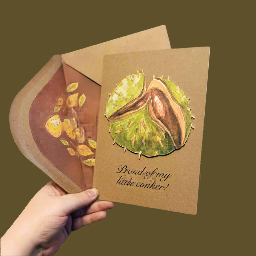 Holding handmade A5 Kraft brown card and envelope. Card has a green split conker mounted in the front and