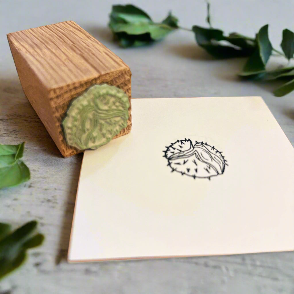 Wooden stamp rests on a black ink conker stamped on a white page. They sit in a rey wooden desk with green foliage.