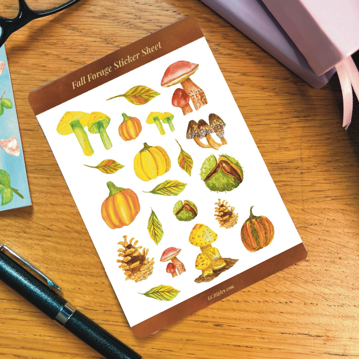 White sticker sheet with 20 colourful mushrooms and autumnal botanicals in a variety of sizes and colours, on a wooden desk with notepad, glasses and a pile of journals