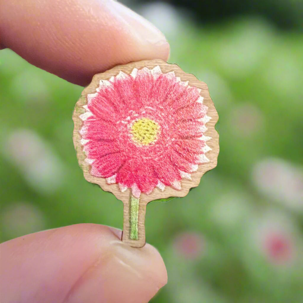 Pink Gerbera wooden pin held in front of a blurred flower garden