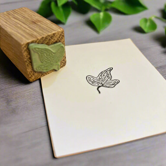 Ivy rubber stamp in green mounted in a wooden block lying next to a sheet of white paper with a black ink stamped. Green foliage decorates the grey wooden desk it sits on