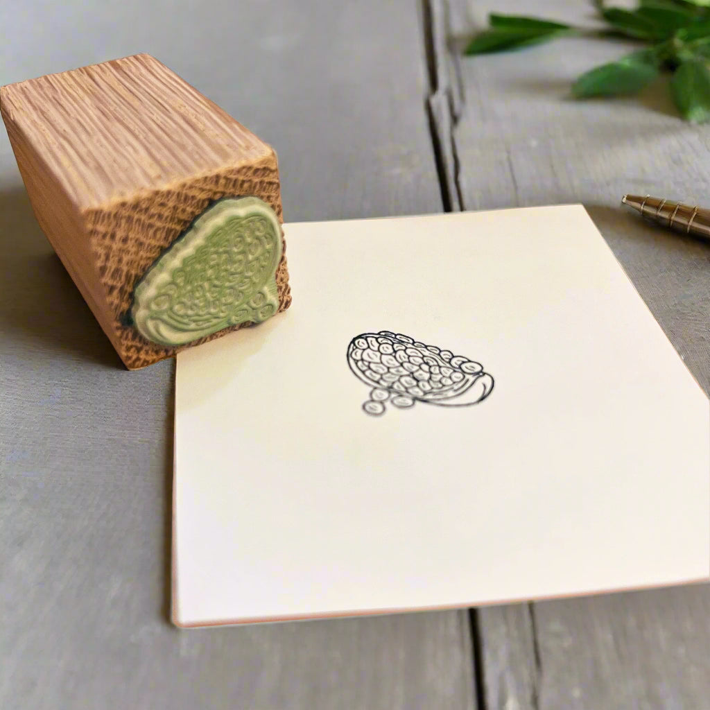 Pomegranate ink stamped on a square of white paper with a wooden block green rubber stamp sat beside. Background is a grey wood stained table with a pen and green foliage,
