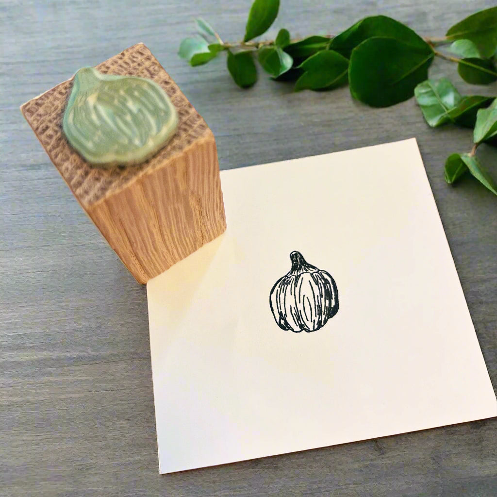 Pumpkin stamped in black ink on white paper with a wooden pumpkin stamp, both on a grey wooden desk with green foliage