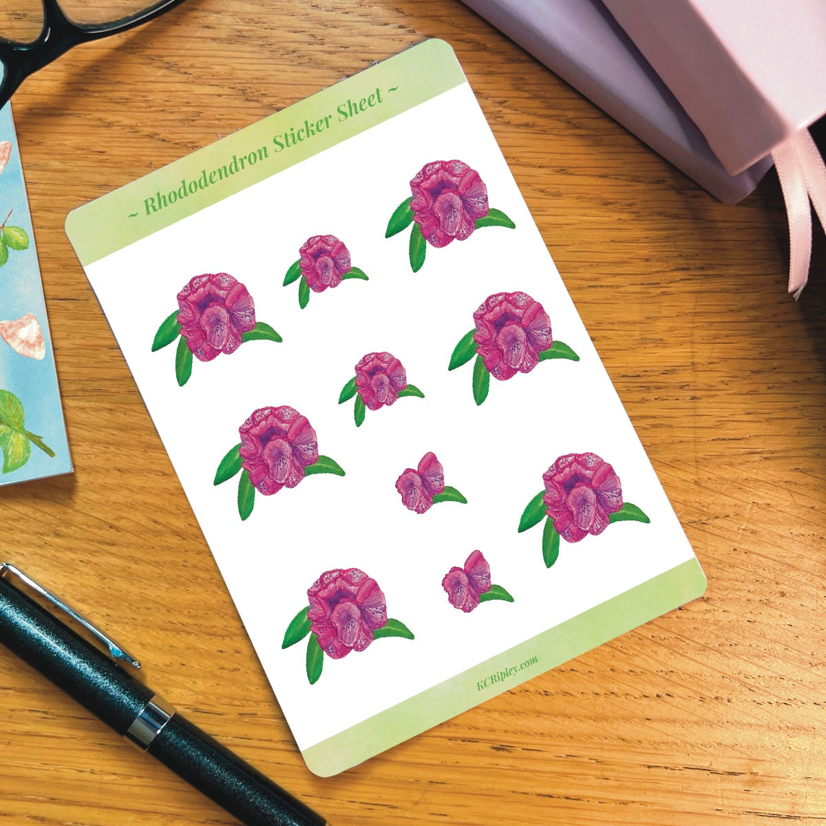 White sticker sheet with 10 pink rhododendron floral stickers, on a wooden desk next to a pile of journals and stationery