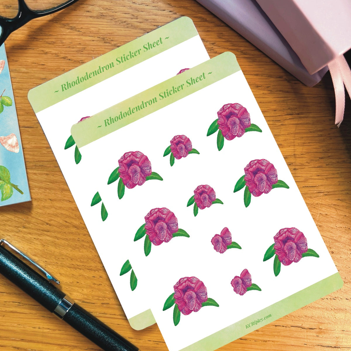 2 White sticker sheets with 10 pink rhododendron floral stickers, on a wooden desk next to a pile of journals and stationery