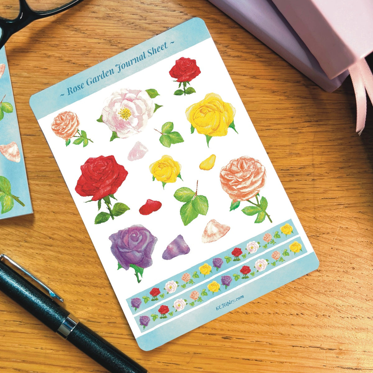 White sticker sheet with 15 pink roses in a variety of sizes and colours and 2 narrow sticker strips in sky blue with repeating rose pattern, on a wooden desk next to a pile of journals and stationery