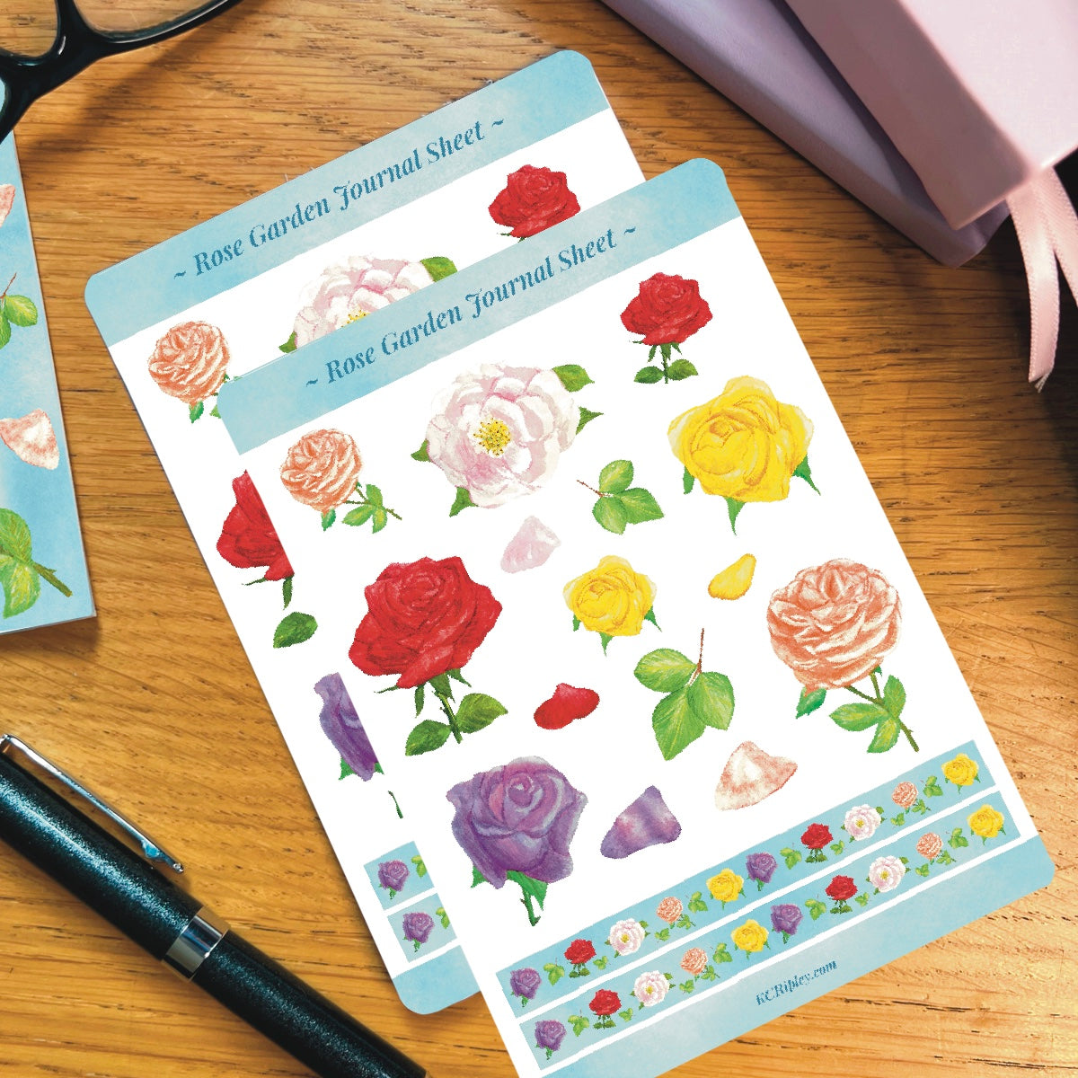 2 White sticker sheets with 15 colourful roses in a variety of sizes and colours and 2 narrow sky blue strips with repeating rose pattern, on a wooden desk next to a pile of journals and stationery