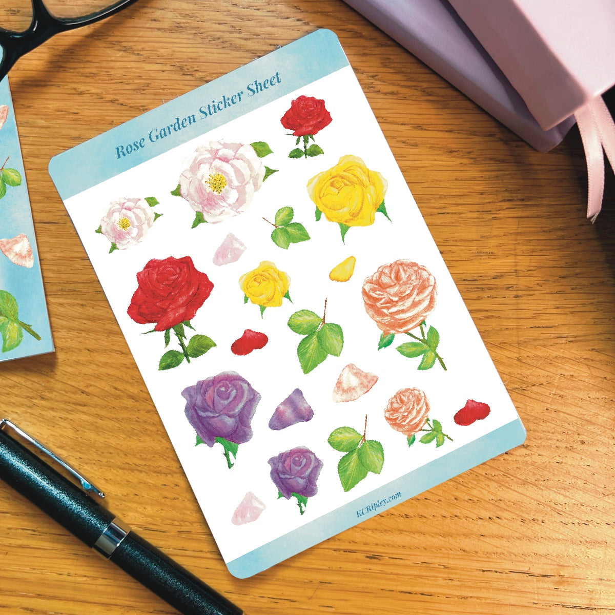 White sticker sheet with 20 colourful roses in a variety of sizes and colours, on a wooden desk next to a pile of journals and stationery