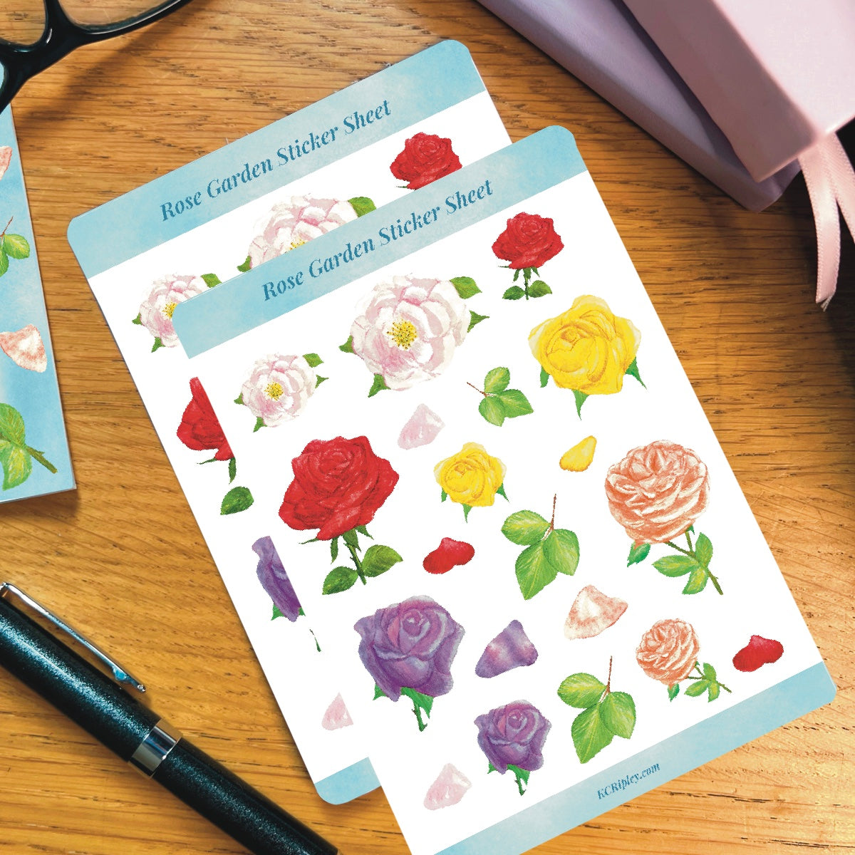 2 White sticker sheets with 20 colourful roses in a variety of sizes and colours, on a wooden desk next to a pile of journals and stationery