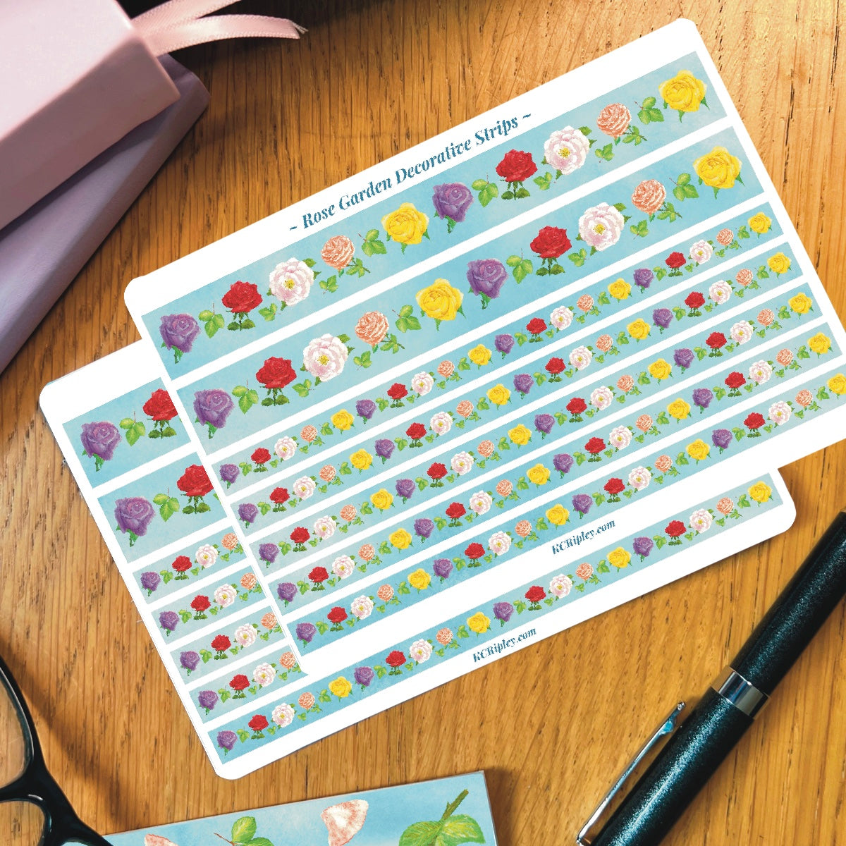 2 White sticker sheets with 2 wide and 2 narrow sticker strips. Strips are light blue with a repeating pattern of colourful roses. Sheets sit on a wooden desk with journals and a pen