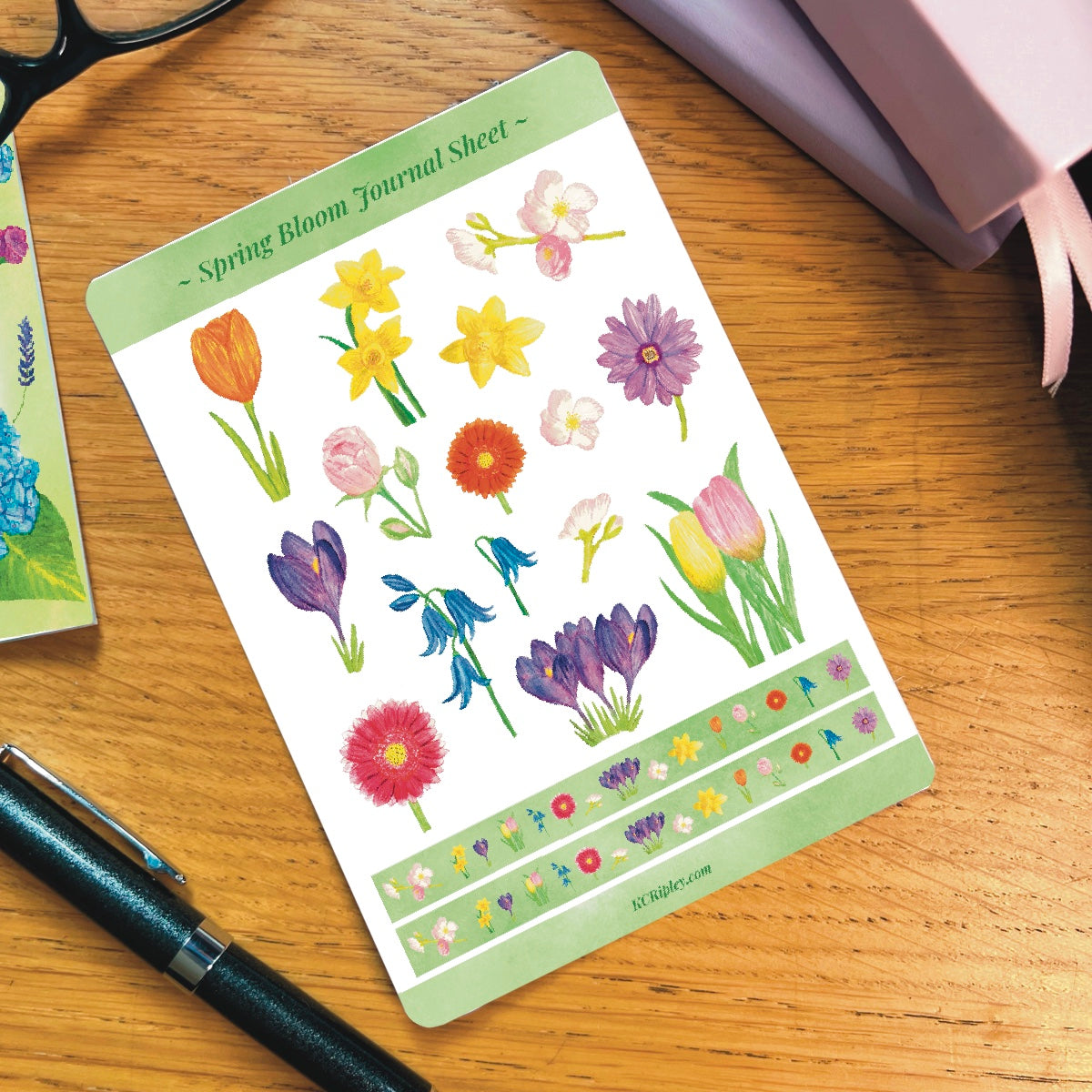 White sticker sheet with 15 colourful spring florals in a variety of sizes and colours and 2 narrow sticker strips in bright green with a repeating floral pattern, on a wooden desk next to a pile of journals and stationery