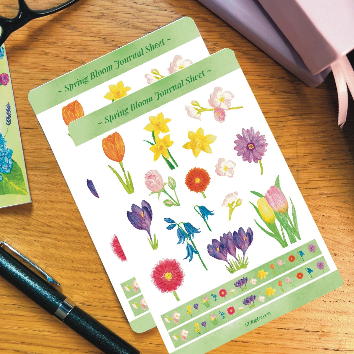 2 White sticker sheet with 15 colourful spring florals in a variety of sizes and colours and 2 narrow sticker strips in bright green with a repeating floral pattern, on a wooden desk next to a pile of journals and stationery