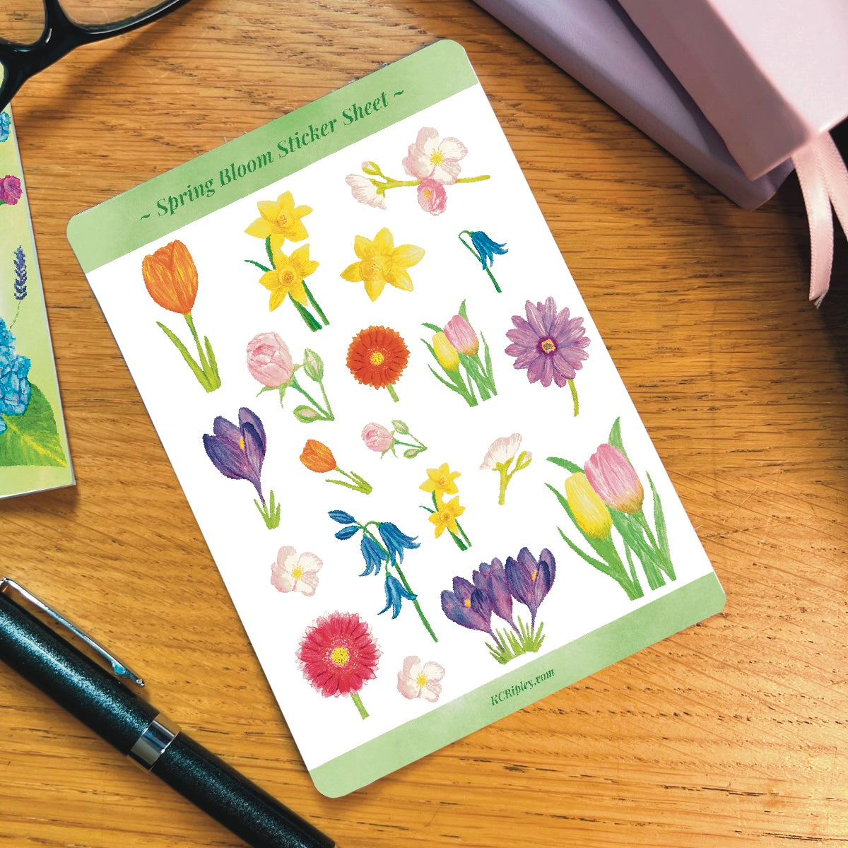 White sticker sheets with 20 spring florals in a variety of sizes, on a wooden desk next to a pile of journals and stationery