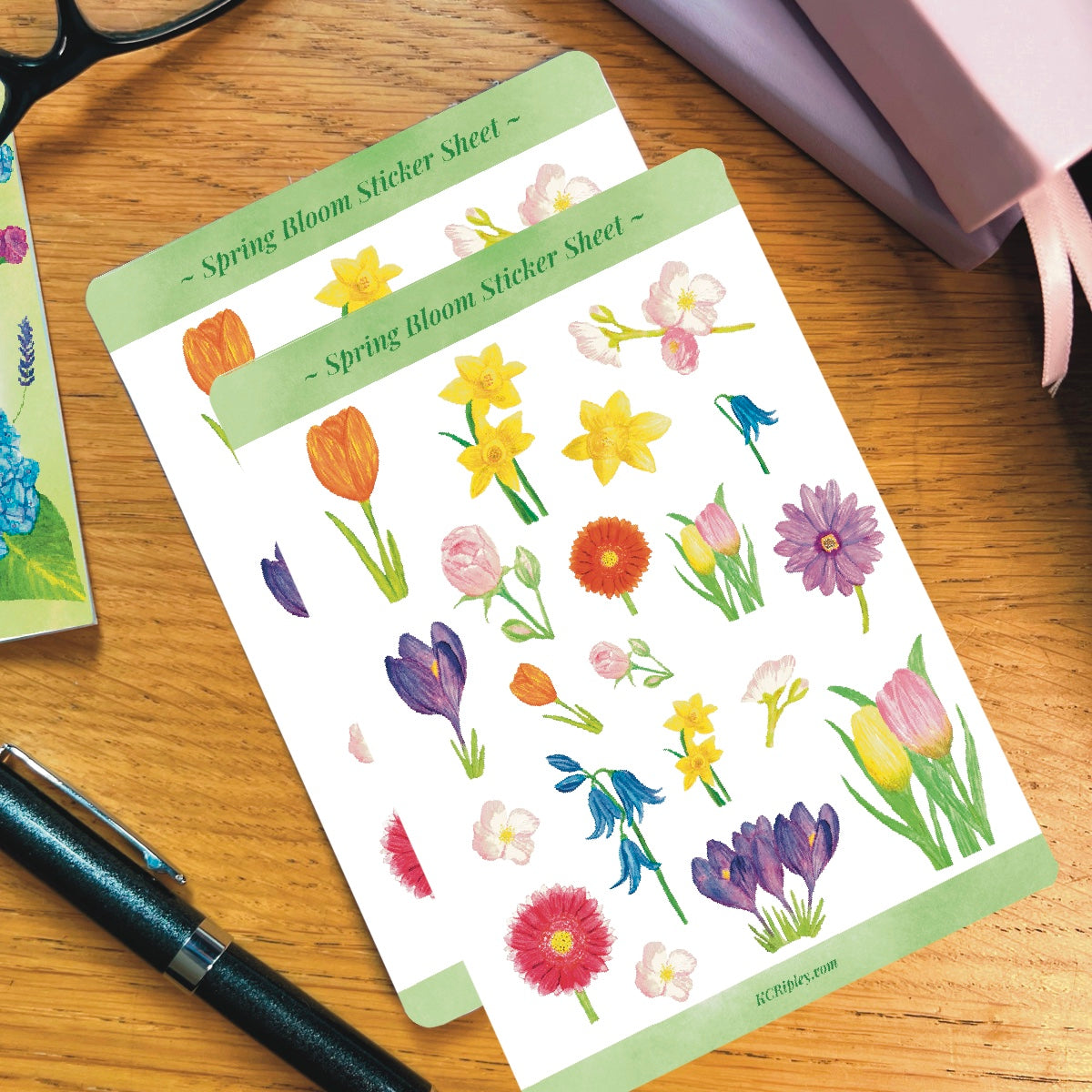 2 White sticker sheets with 20 spring florals in a variety of sizes, on a wooden desk next to a pile of journals and stationery