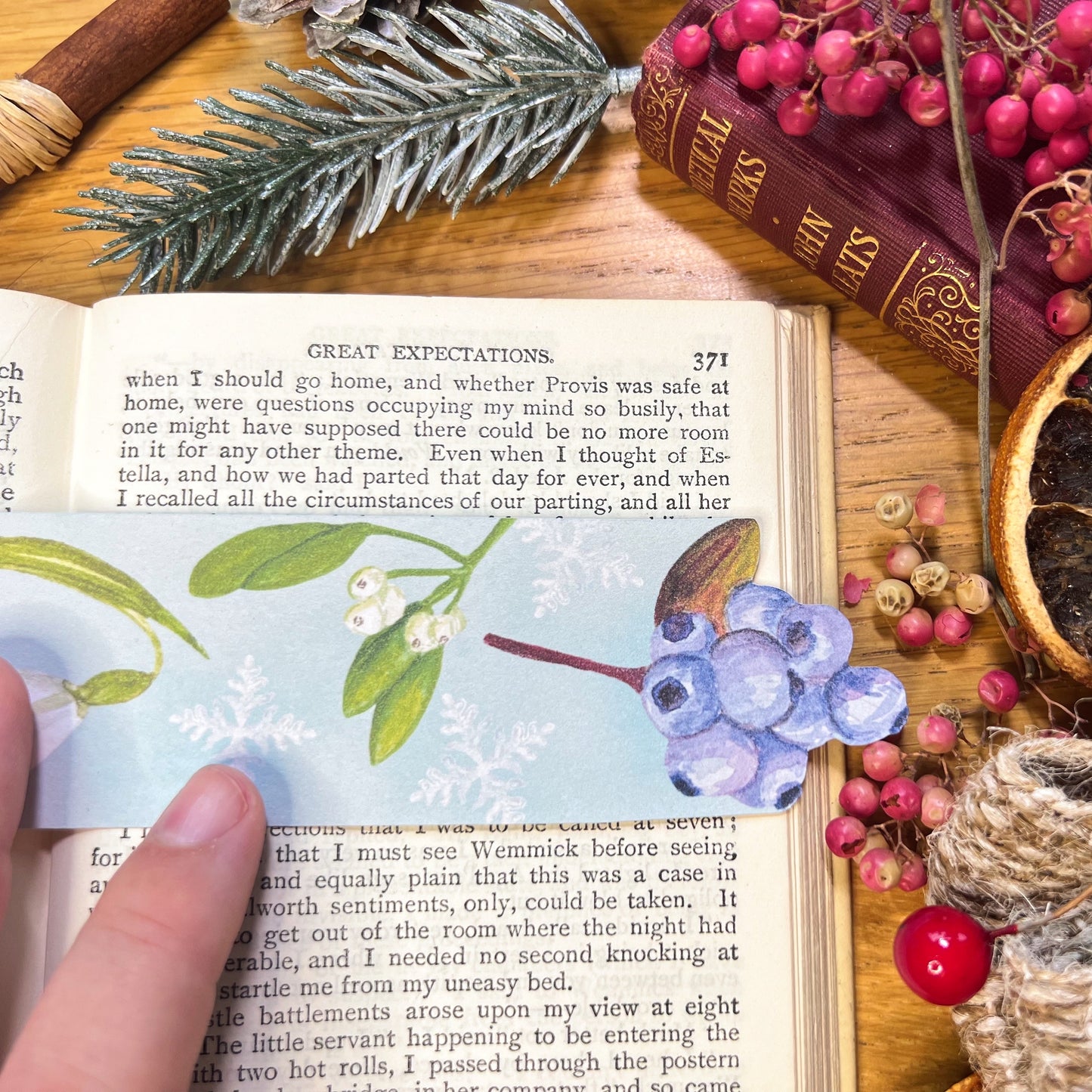 Close up of winter wonderland illustrated bookmark used to read open pages of a book, cut to the shape of blueberries