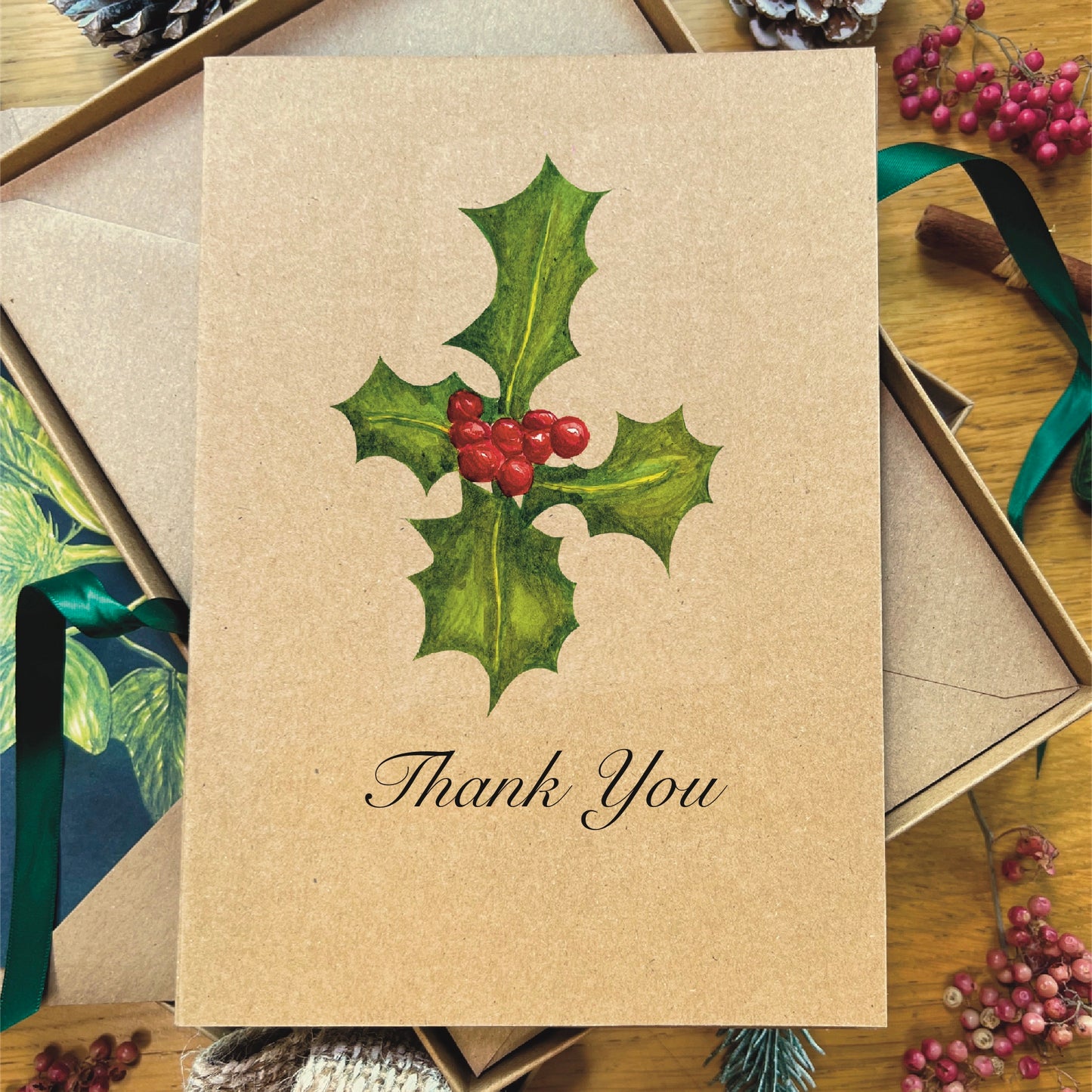 English Holly sprig and berries painted in watercolour attached to a natural recycled blank greetings card, laying in a box with an illustrated inlay envelope
