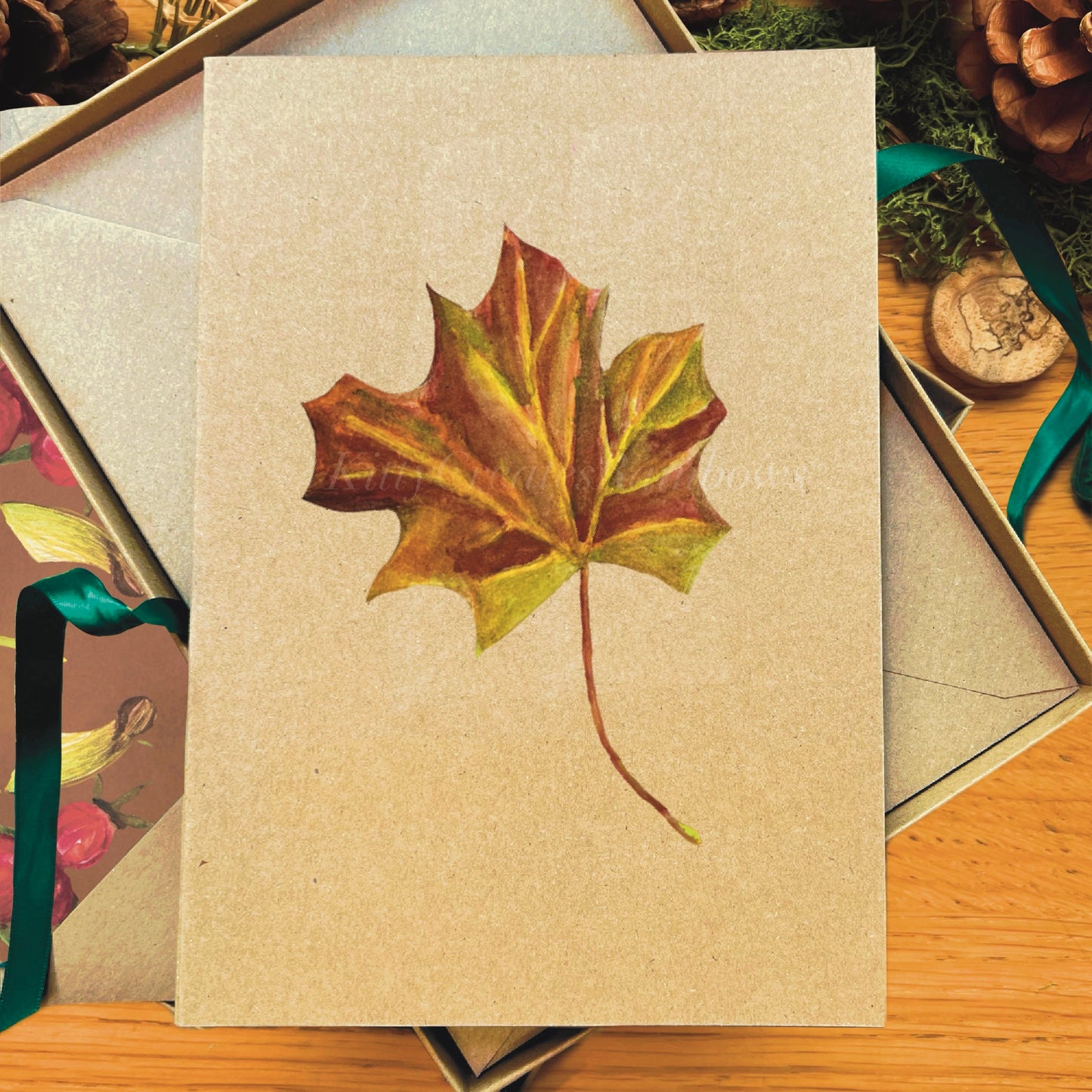 Maple leaf painted in watercolour attached to a natural recycled blank greetings card, laying in a box with an illustrated inlay envelope