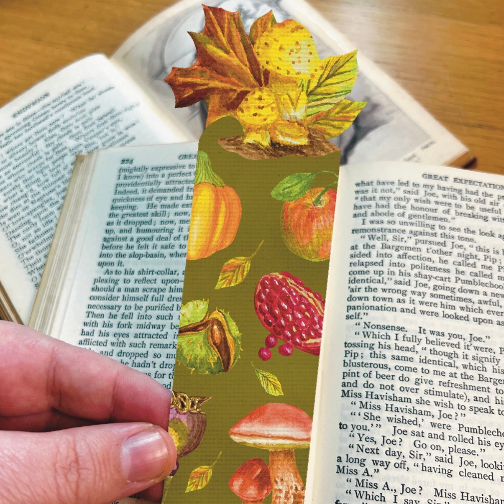 Bookmark in moss green illustrated with harvest fruits and mushrooms, die cut to the shape of maple leaf and mushrooms.  Bookmark is held ion front of the open pages of a book.