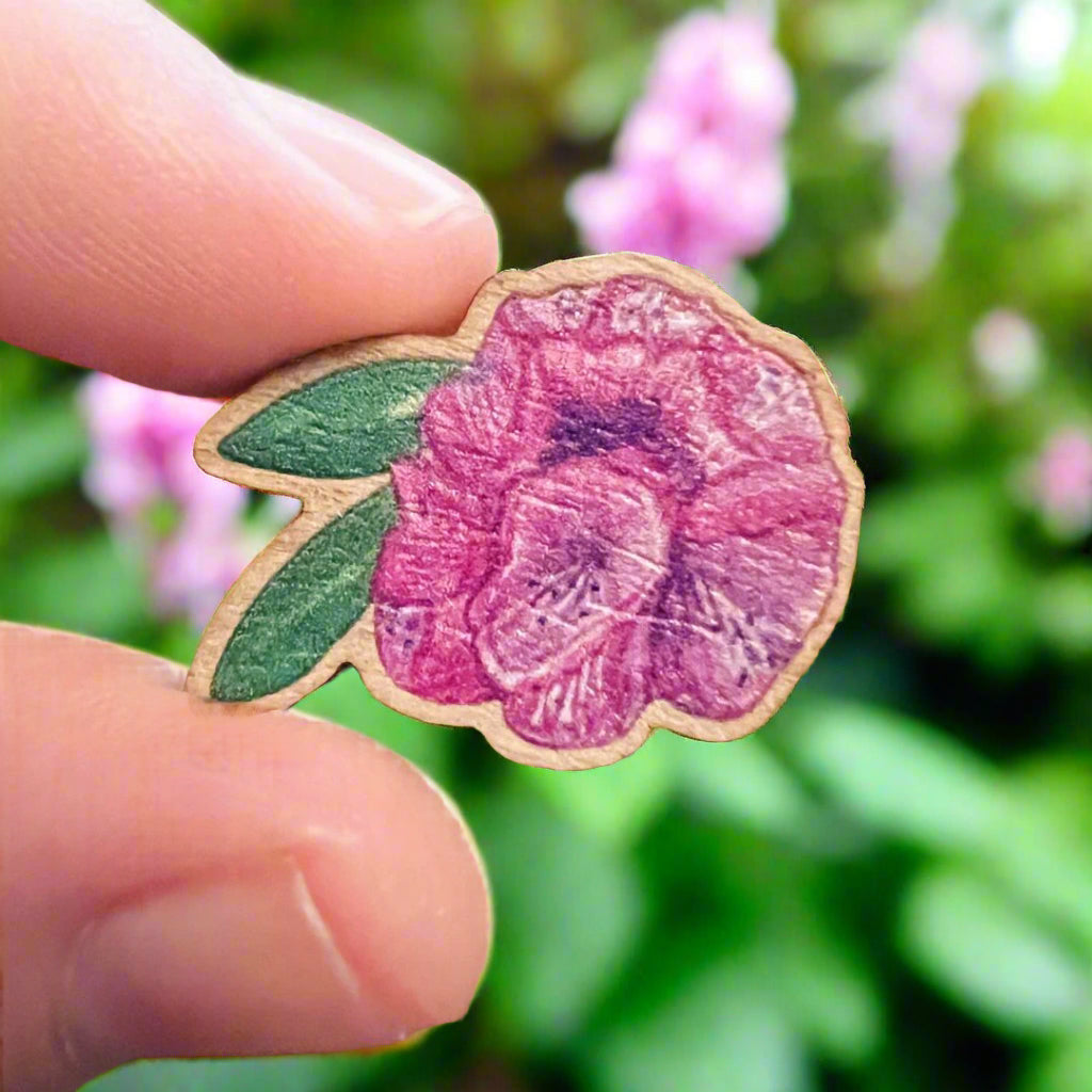 Pink rhododendron die cut wooden pin held in front of a pink and green flower bed blurred in the background 