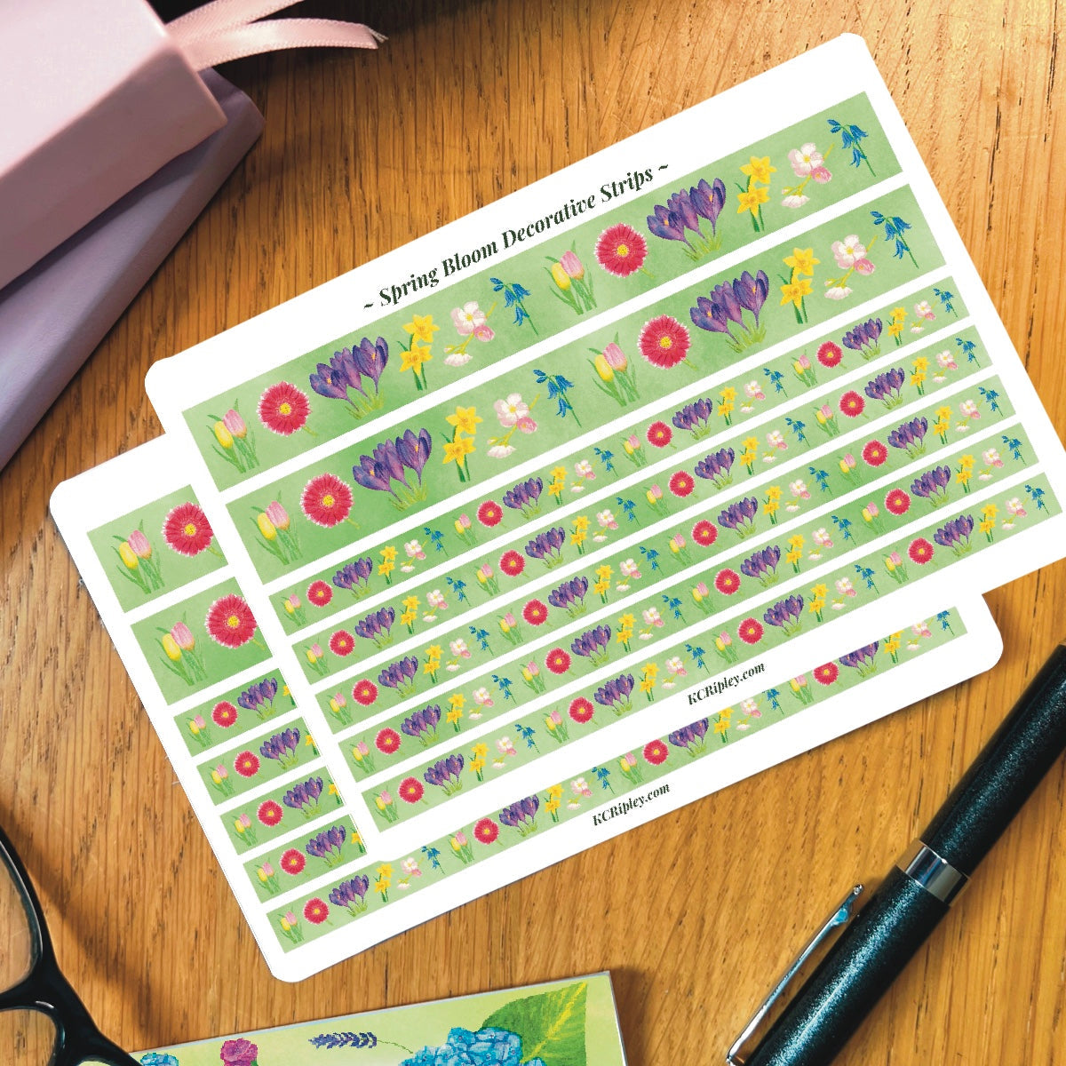 2 White sticker sheet with 2 wide and 5 narrow decorative sticker strips of colourful spring florals with a bright green background in a repeating floral pattern, on a wooden desk next to a pile of journals and stationery