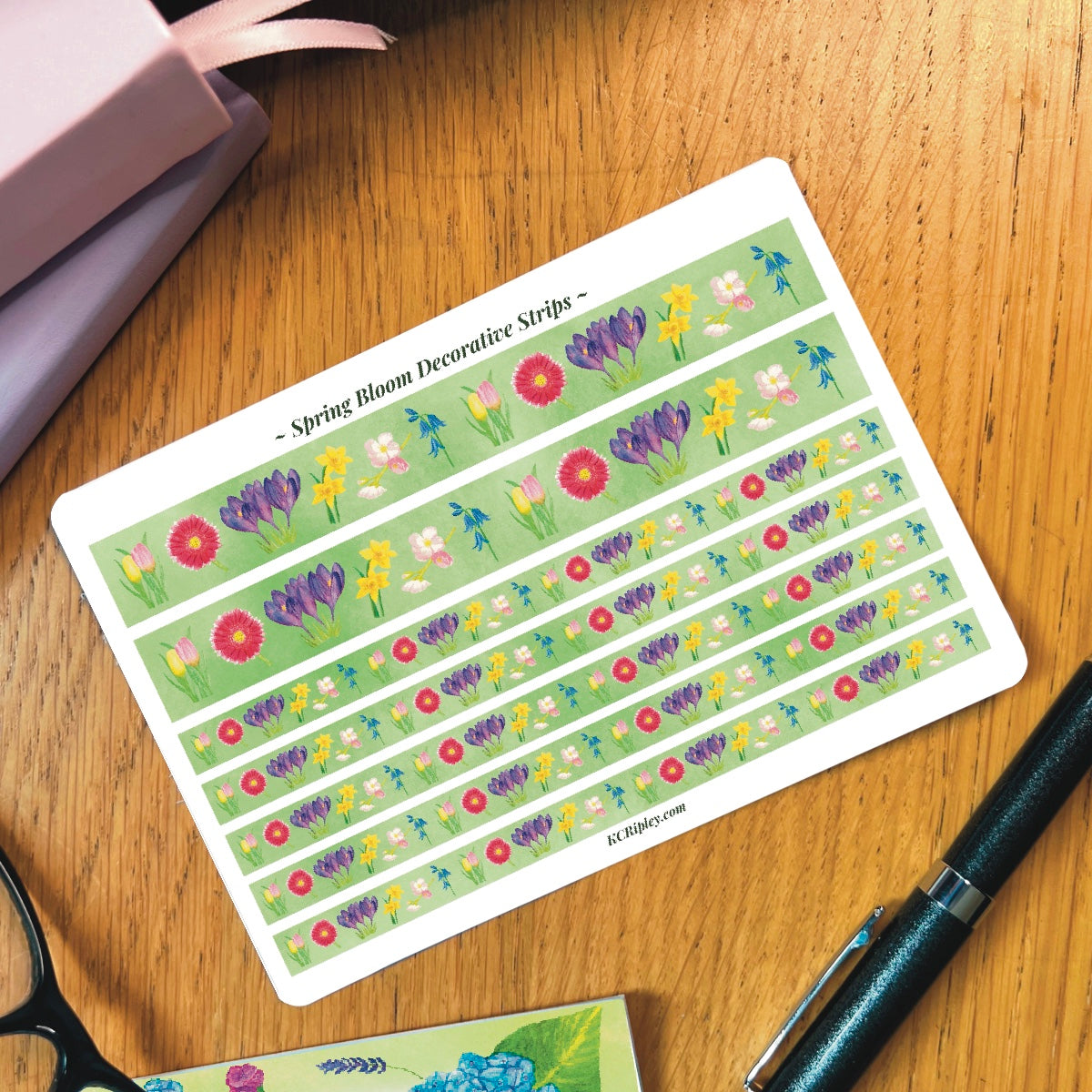 White sticker sheet with 2 wide and 5 narrow decorative sticker strips of colourful spring florals with a bright green background in a repeating floral pattern, on a wooden desk next to a pile of journals and stationery