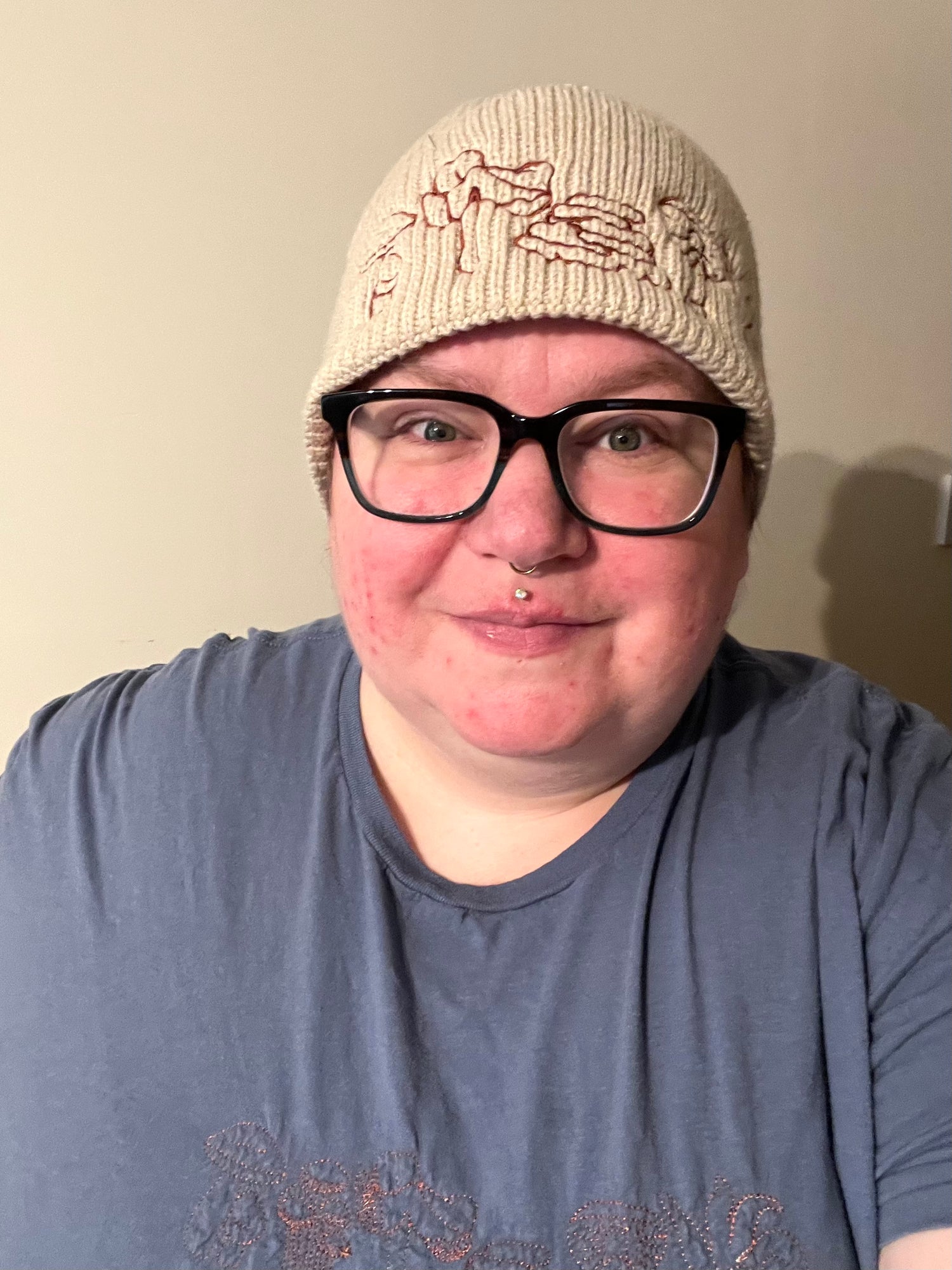 Ripley a plus size person wears a cream woollen beanie with mushrooms embroidered on the cuff. They’re smiling wearing a blue T-shirt and black rimmed glasses